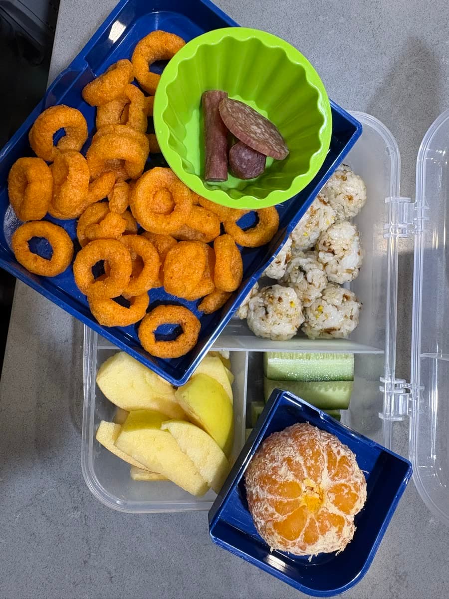 A real packed Kiwi Lunchbox: popcorn bites, cucumber sticks, peeled mandarin, apple slices, salami in a silicone cup, and ring snacks in a blue bento box